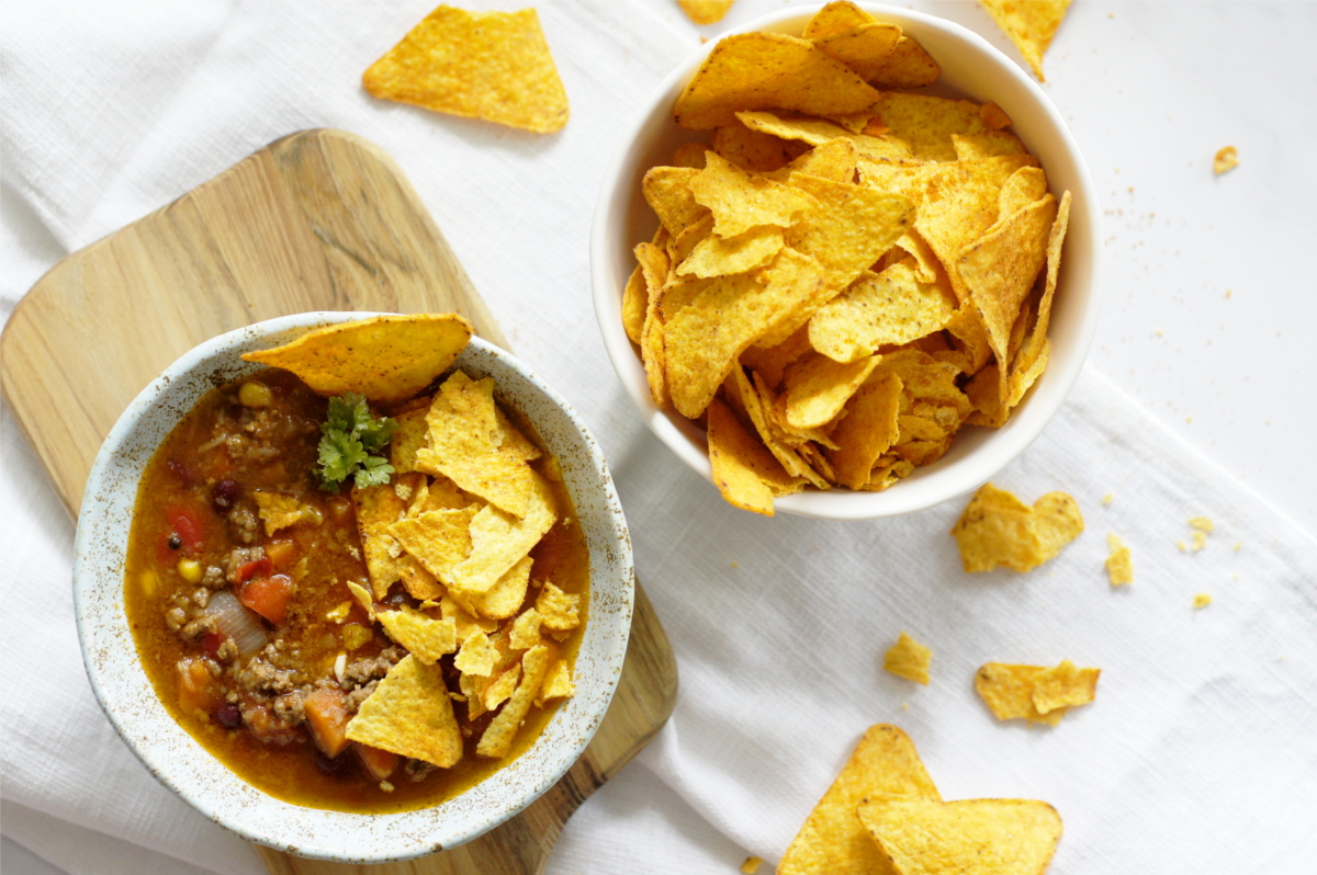 W ceramicznej miseczce jest zupa chili con carne z pokruszonymi chipsami tortilla. Miseczka stoi na drewnianej deseczce na stole.  Obok stoi miska z porcją chipsów tortilla. Dookoła leżą kawałki chipsów. 