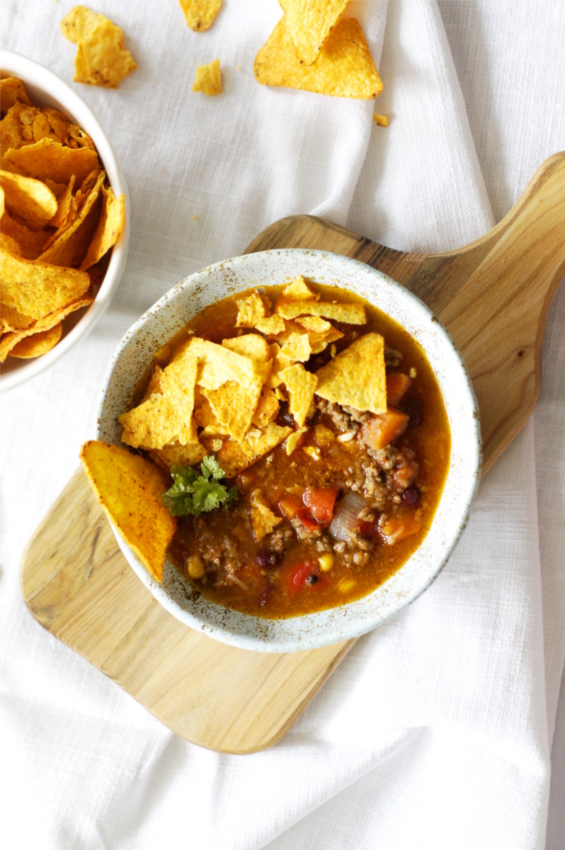 W ceramicznej miseczce jest zupa chili con carne z pokruszonymi chipsami tortilla. Miseczka stoi na drewnianej deseczce na stole.  Obok stoi miska z porcją chipsów tortilla. Dookoła leżą kawałki chipsów. 