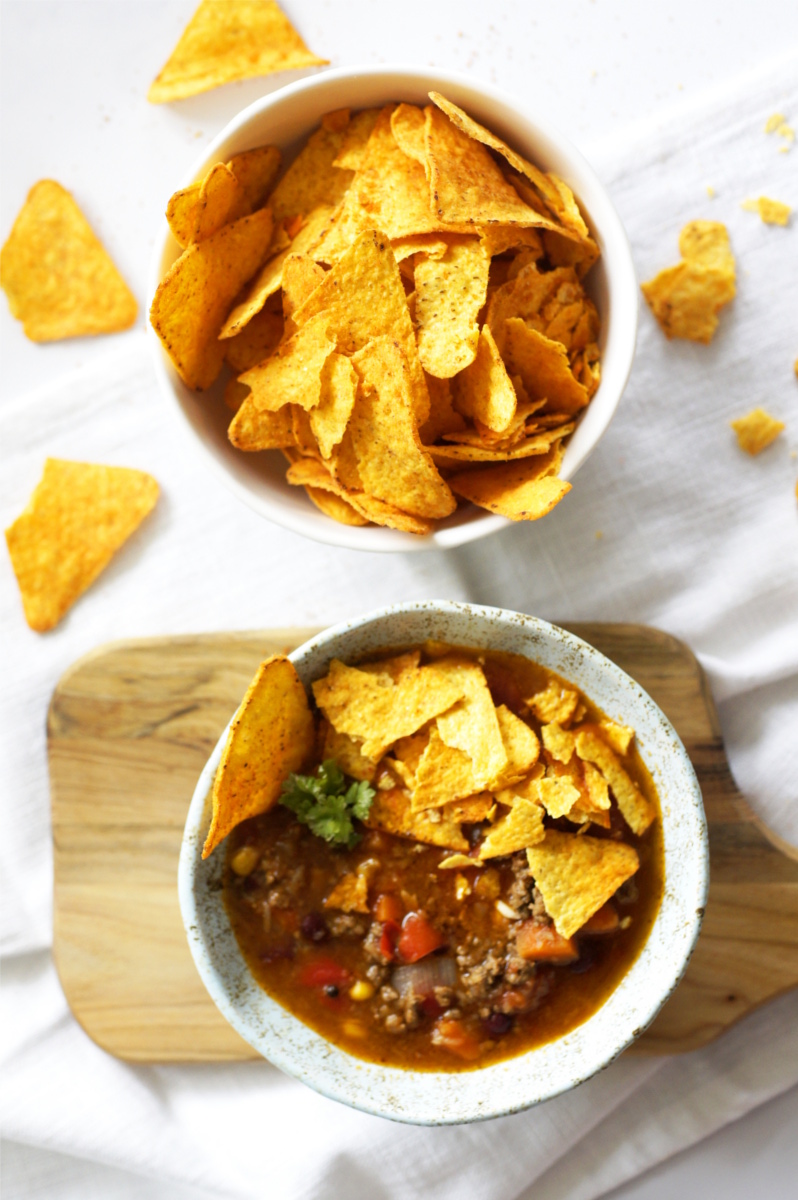 W ceramicznej miseczce jest zupa chili con carne z pokruszonymi chipsami tortilla. Miseczka stoi na drewnianej deseczce na stole.  Obok stoi miska z porcją chipsów tortilla. Dookoła leżą kawałki chipsów. 