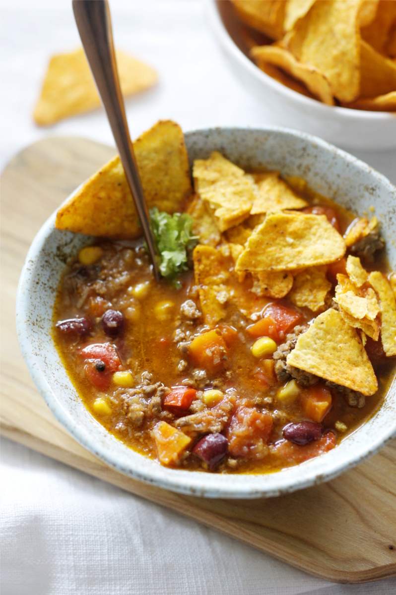 W ceramicznej miseczce jest zupa chili con carne z pokruszonymi chipsami tortilla. Miseczka stoi na drewnianej deseczce na stole.  Obok stoi miska z porcją chipsów tortilla.