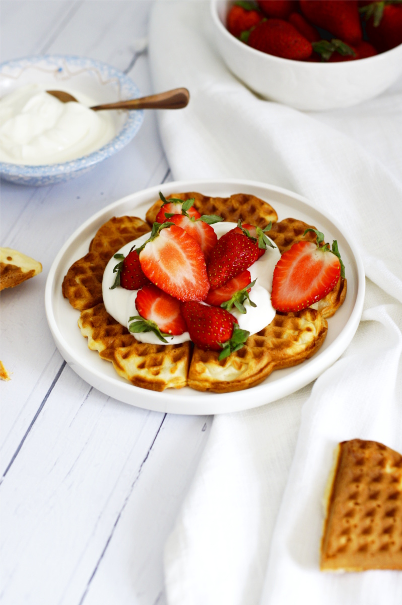 Gofry z dodatkiem delikatnego serka ricotta podane z jogurtem i świeżymi truskawkami. Gofry leżą na białym talerzu.