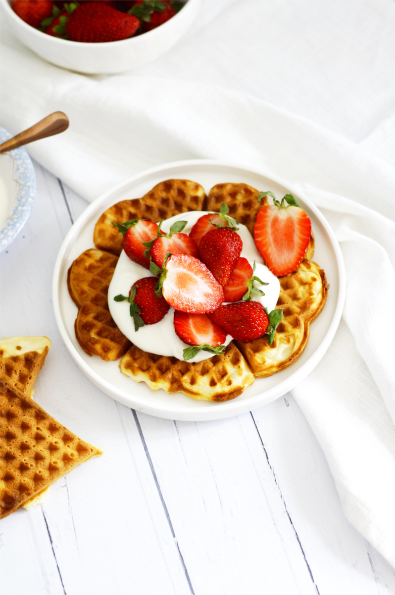 Gofry z dodatkiem delikatnego serka ricotta podane z jogurtem i świeżymi truskawkami. Gofry leżą na białym talerzu.